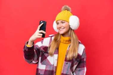 Young beautiful woman wearing winter muffs isolated on red background making a selfie