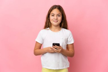 Child over isolated pink background looking at the camera and smiling while using the mobile