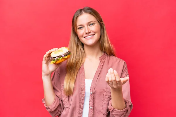 Young caucasian woman holding a burger  isolated on red background inviting to come with hand. Happy that you came