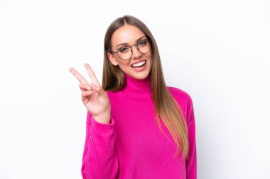 Beyaz arka planda izole edilmiş genç beyaz kadın gülümsüyor ve zafer işareti gösteriyor.