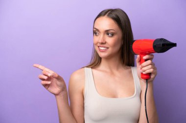 Young caucasian woman holding a hairdryer isolated on purple background pointing to the side to present a product