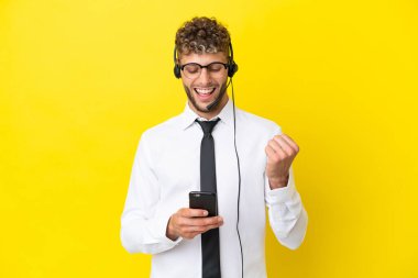 Telemarketer blonde man working with a headset isolated on yellow background with phone in victory position