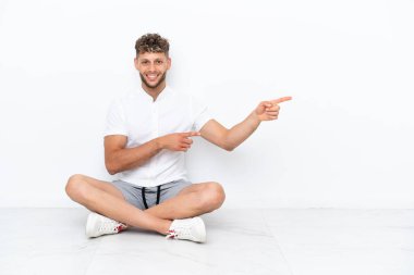 Young blonde man sitting on the floor isolated on white background pointing finger to the side and presenting a product