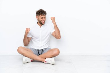 Genç sarışın adam, beyaz arka planda izole bir şekilde yerde oturmuş zaferi kutluyor.