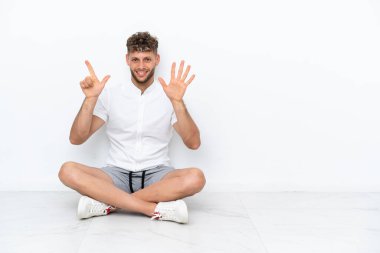 Young blonde man sitting on the floor isolated on white background counting seven with fingers
