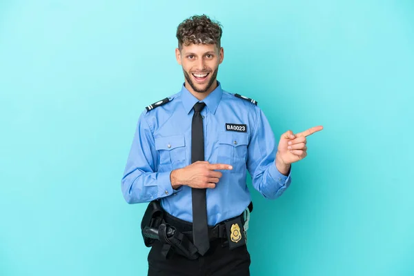 Young police blonde man isolated white on blue background surprised and pointing side