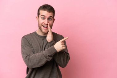 Pembe arka planda izole edilmiş genç beyaz bir adam bir ürün sunmak ve bir şeyler fısıldamak için kenarı işaret ediyor.