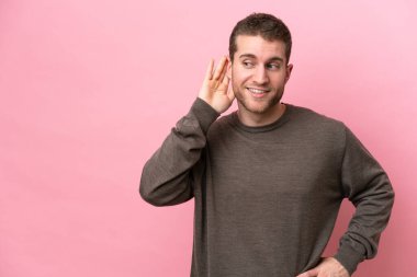 Pembe arka planda izole edilmiş genç beyaz adam elini kulağına koyarak bir şeyler dinliyor.