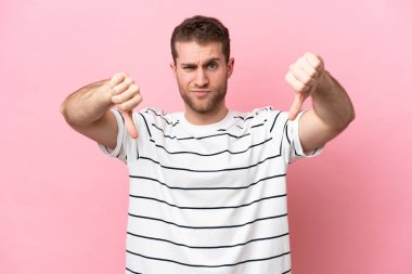 Pembe arka planda izole edilmiş genç beyaz adam baş parmağını iki eliyle aşağı gösteriyor.