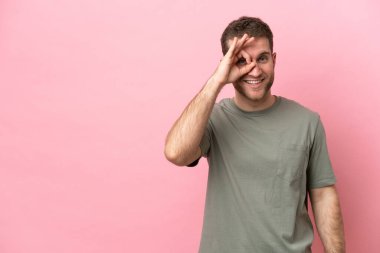 Pembe arka planda izole edilmiş genç beyaz adam parmaklarıyla tamam işareti gösteriyor.