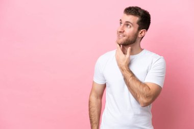 Pembe arka planda izole edilmiş genç beyaz adam gülümserken yukarı bakıyor.