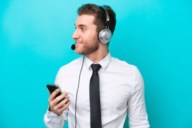 Telemarketer caucasian man working with a headset isolated on blue background keeping a conversation with the mobile phone with someone