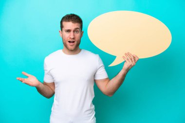 Kafkasyalı genç adam mavi arka planda izole edilmiş. Elinde şaşırmış bir ifadeyle boş bir konuşma balonu tutuyor.