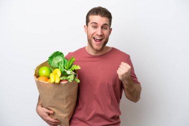 Young caucasian man holding a grocery shopping bag isolated on white background celebrating a victory in winner position