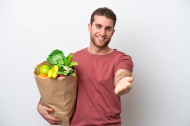 Young caucasian man holding a grocery shopping bag isolated on white background shaking hands for closing a good deal