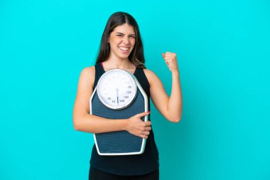 Young Italian woman isolated on blue background with weighing machine and doing victory gesture