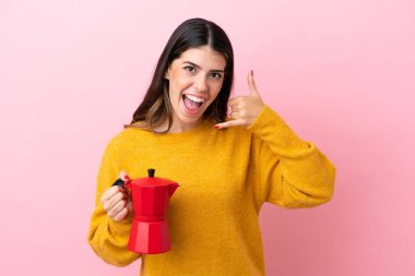 Young Italian woman holding a coffee maker isolated on pink background making phone gesture. Call me back sign