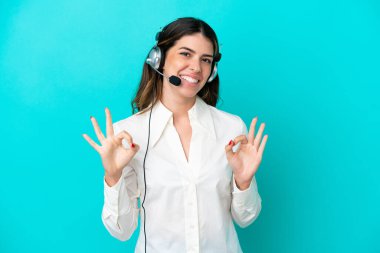 Telemarketer Italian woman working with a headset isolated on blue background showing an ok sign with fingers