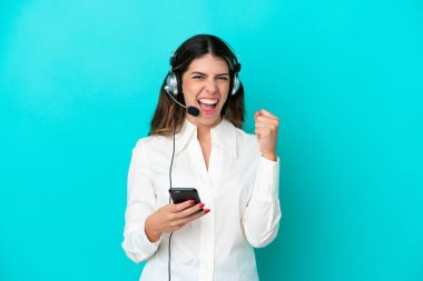 Telemarketer Italian woman working with a headset isolated on blue background with phone in victory position