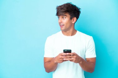 Kafkasyalı genç adam mavi arka planda izole edildi cep telefonu kullanarak ve yukarı bakarak