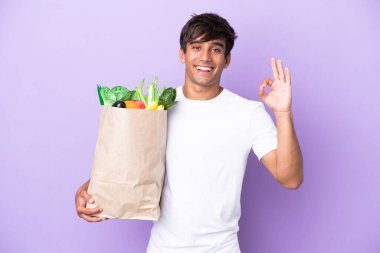 Young man holding a grocery shopping bag isolated on purple background showing ok sign with fingers