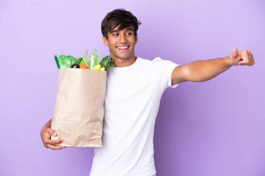 Young man holding a grocery shopping bag isolated on purple background giving a thumbs up gesture