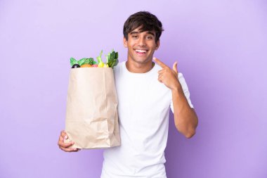 Young man holding a grocery shopping bag isolated on purple background giving a thumbs up gesture