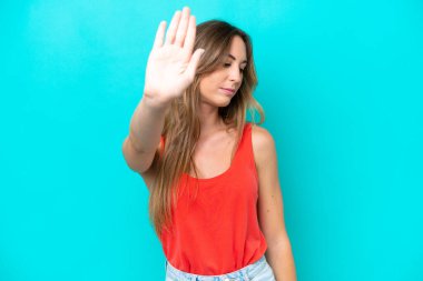 Mavi arka planda izole edilmiş genç beyaz kadın dur hareketi yapıyor ve hayal kırıklığına uğruyor.