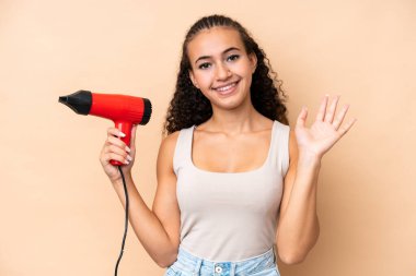 Young woman holding a hairdryer isolated on beige background saluting with hand with happy expression