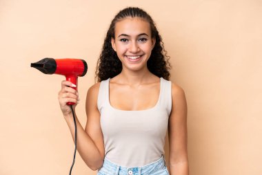 Young woman holding a hairdryer isolated on beige background smiling a lot