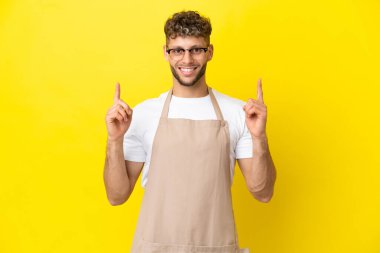 Restoran garsonu sarı arka planda izole edilmiş sarışın adam harika bir fikre işaret ediyor.