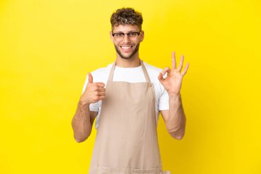 Restoran garsonu. Sarı arka planda izole edilmiş sarışın adam. Tamam işareti gösteriyor.