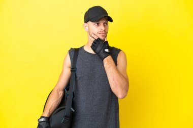 Spor çantalı genç sarışın adam sarı arka planda izole edilmiş. Şüpheleri var ve kafası karışık.