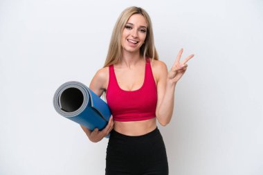 Genç sarışın spor kadın yoga derslerine gidiyor beyaz arka planda izole edilmiş bir minder tutarken gülümsüyor ve zafer işareti gösteriyor.