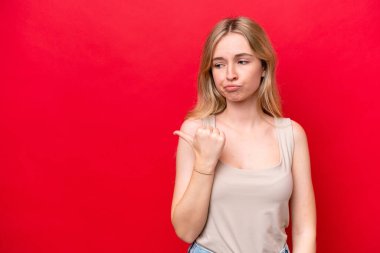 Young English woman isolated on red background unhappy and pointing to the side