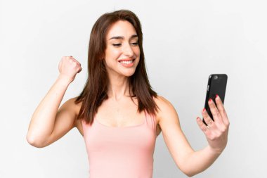 Beyaz arka plan üzerinden izole edilmiş genç beyaz kadın cep telefonu kullanıyor ve zafer jesti yapıyor.