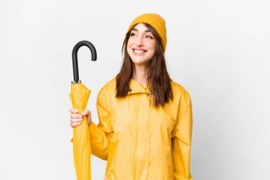 Young caucasian woman with rainproof coat and umbrella over isolated white background thinking an idea while looking up