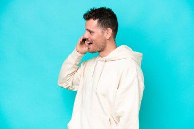 Beyaz tenli yakışıklı adam mavi arka planda izole edilmiş cep telefonuyla biriyle sohbet ediyor.