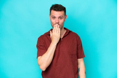 Beyaz tenli yakışıklı bir adam mavi arka planda izole edilmiş. Ağzını eliyle kapatıyor.