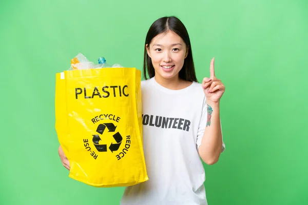 Young Asian woman holding a bag full of plastic bottles to recycle over isolated chroma key background pointing up a great idea
