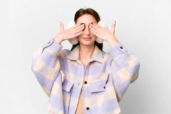 Young caucasian woman wearing winter muffs over isolated white background covering eyes by hands