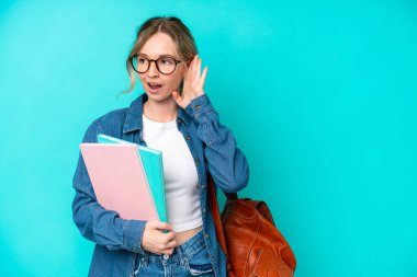 Genç bayan öğrenci, mavi arka planda izole edilmiş. Kulağına elini sürerek bir şeyler dinliyor.