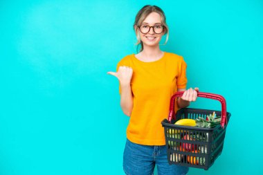 Blonde English young girl holding a shopping basket full of food isolated on blue background pointing to the side to present a product