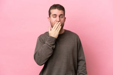 Pembe arka planda izole edilmiş genç beyaz adam esniyor ve ağzını eliyle açık tutuyor.