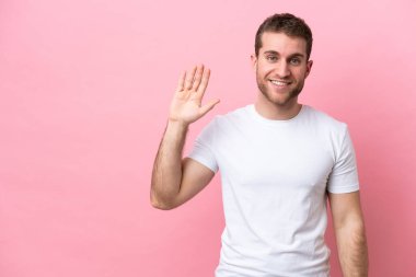 Pembe arka planda izole edilmiş genç beyaz adam mutlu bir ifadeyle selamlıyor.