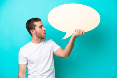 Mavi arka planda izole genç beyaz adam boş bir konuşma balonu tutuyor.