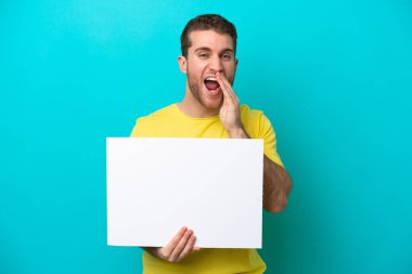 Young caucasian man isolated on blue background holding an empty placard and shouting