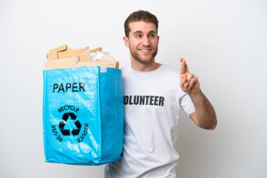 Young caucasian man holding a recycling bag full of paper to recycle isolated on white background with fingers crossing and wishing the best