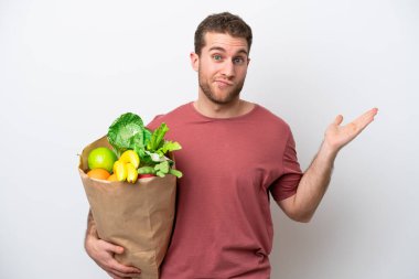 Young caucasian man holding a grocery shopping bag isolated on white background having doubts while raising hands