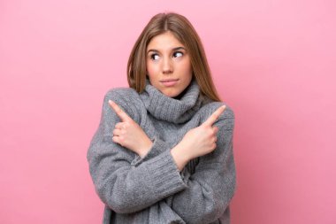 Young caucasian woman isolated on pink bakcground pointing to the laterals having doubts